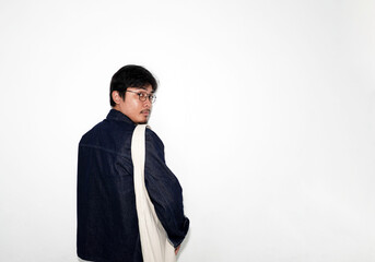 Asian man in dark denim jacket, glasses, and tote bag, looking back over his shoulder with a serious or inquisitive expression against a white background with copy space.