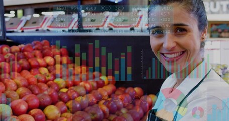Smiling clerk in apron tracking financial charts over red apple bin in produce section, copy space