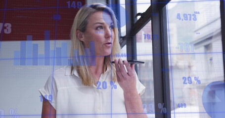 Dictating businesswoman wearing white blouse using smartphone at office window, with chart overlays