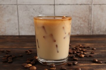 Tasty iced coffee with milk in glass and beans on wooden table against tiled wall, closeup