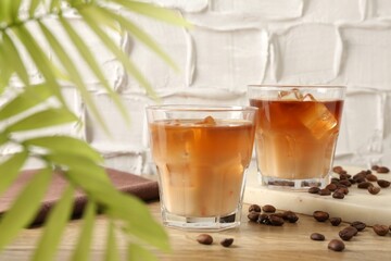 Tasty iced coffee with milk in glasses, beans and leaves on wooden table against light textured background, closeup