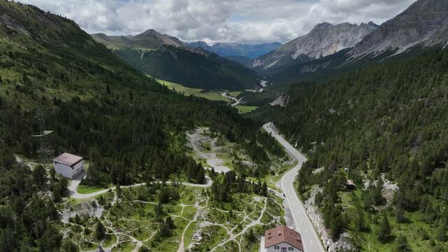 Aerial view of a winding road cutting through lush green mountains, contrasting with the rugged peaks, Val Mustair, Grisons, Switzerland.