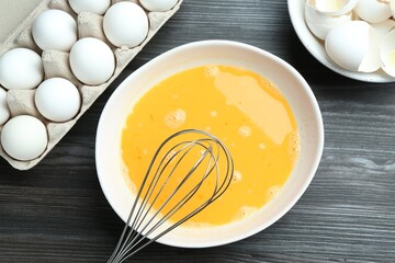 Beaten eggs in bowl and whisk on dark wooden table, flat lay