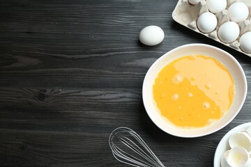 Beaten eggs in bowl and whisk on dark wooden table, flat lay. Space for text