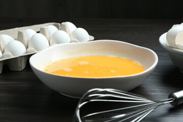 Beaten eggs in bowl and whisk on dark wooden table, closeup
