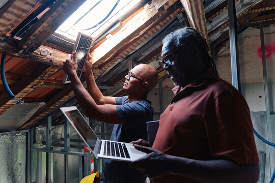 Senior architect using laptop with builder examining solar panel by window at site