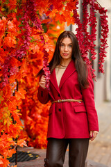 Portrait of a young woman with leaves in an autumn park. A stylish girl in a burgundy jacket strolls outdoors. Autumn fashion.
