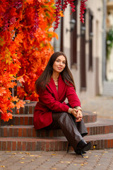 Portrait of a young woman with leaves in an autumn park. A stylish girl in a burgundy jacket strolls outdoors. Autumn fashion.