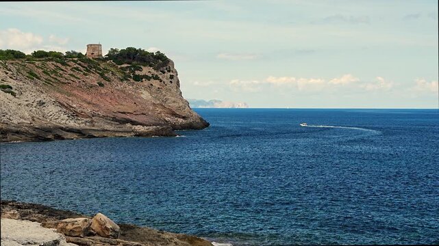 Motorboat cruising near rocky cliffs with Torre d Albarca tower on Mallorca, Spain. Clear blue sea and scenic Mediterranean coastal landscape under sunny sky.