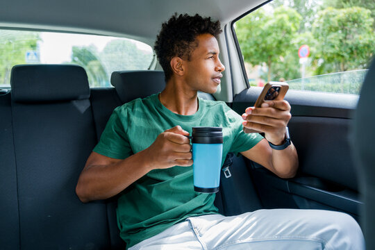 Man in backseat of car holding coffee and smartphone during commute