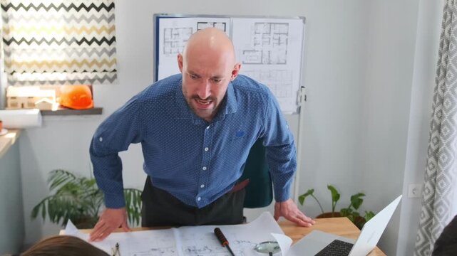 Bald architect angrily leans over desk and scolds employees for a mistake in construction blueprints. Intense office scene with engineers. Team looks stressed as they try to correct the problem.