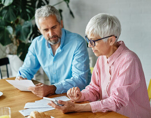 Senior couple checking bills and calculating expenses using a calculator at home, debt taxes and...