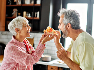 Portrait of happy senior couple prepering meal and tasting pizza in kitchen