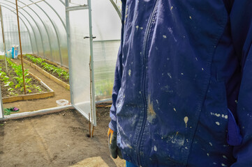 A man stands near a greenhouse in dirty clothes.