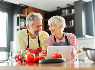 Portrait of happy senior mid aged mature couple prepering meal with fresh vegatebles and following internet instructions for a recipe on a tablet computer  or looking at video  or website app in kitch