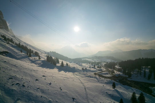Aerial view of a crisp, sun-drenched alpine landscape where snow-laden slopes meet a hazy horizon, cradling a valley dotted with evergreens and the distant promise of mountain peaks, Switzerland.