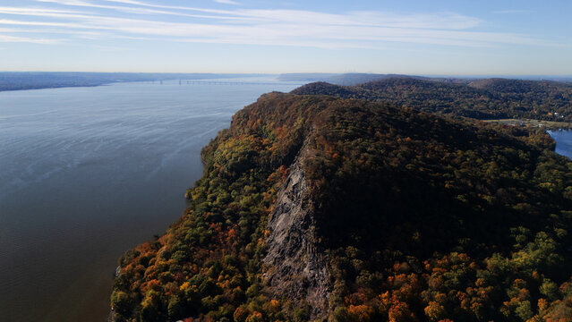Aerial view of a dramatic cliff face plunging into the Hudson River, adorned with autumnal foliage, creating a tapestry of warm hues against the cool water, Nyack, New York, United States.