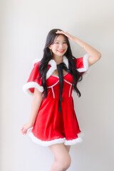 A young woman wearing a red santa dress with white fur trim and a black ribbon poses cheerfully against a plain white wall, smiling with festive charm in a bright indoor setting.