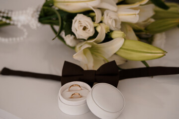Elegant Wedding Rings and Bouquet: Close-up of Gold Bands, White Lilies, Brown Bow Tie on White Fabric, Romantic Celebration Style.