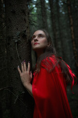 In a mystical forest at twilight, a woman dressed in a flowing red cloak leans against a tree. Her...