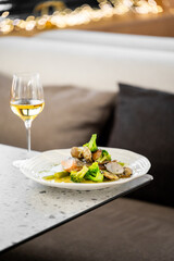 A plate of gourmet seafood with broccoli and clams, accompanied by a glass of white wine, served on a modern speckled table in a restaurant setting