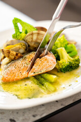 A close-up of a perfectly cooked salmon fillet on a white plate, garnished with steamed broccoli, asparagus, and clams. A fork and knife are cutting into the fish, creating a sense of a fine dining