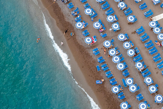Aerial view of azure waters meeting the dark sandy beach dotted with blue-and-white umbrellas and sunbeds, a vibrant contrast of summer hues, Positano, Campania, Italy.