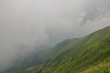 pendii erbosi di un monte, visto dalla cima, che si estendono verso la valle, con un cielo e un ambiente coperto da nuvole e dalla foschia, di giorno, in estate