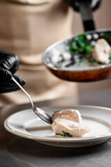 A chef in black gloves uses a spoon to carefully place a portion of cooked meat on a white plate, with a frying pan of food visible in the background