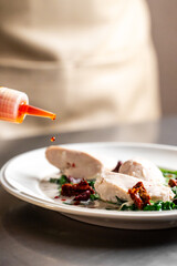 A chef preparing a dish of sliced chicken breast with sun-dried tomatoes and spinach in a creamy sauce, with red oil being drizzled from a bottle