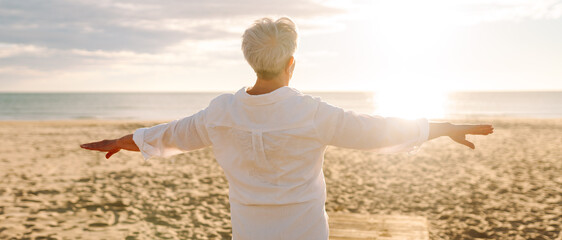 An older woman with gray hair walks along a sunlit sandy beach. The elderly woman enjoys the seascape, feeling the freedom of dawn. Concept of a happy retirement, nature.
