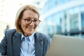 A woman in a suit is sitting at a table with a laptop and a cup of coffee. She is smiling and she is enjoying her work