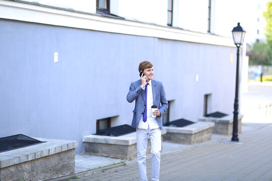 Young man in a stylish suit is walking on a city street while talking on his smartphone and holding a coffee cup, showcasing modern urban lifestyle and professionalism