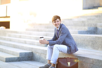 Young man in a stylish suit sitting on outdoor steps, holding a coffee cup, with a leather...