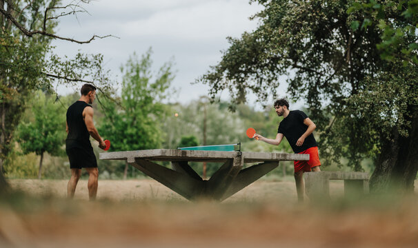 Two men in athletic wear compete in a casual outdoor table tennis match on a park table, smiling and focused as they hit shots under leafy trees.