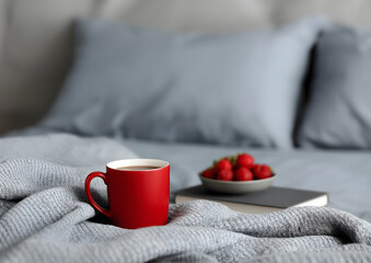 Cozy red mug and bowl of strawberries on bed with gray blanket and soft light