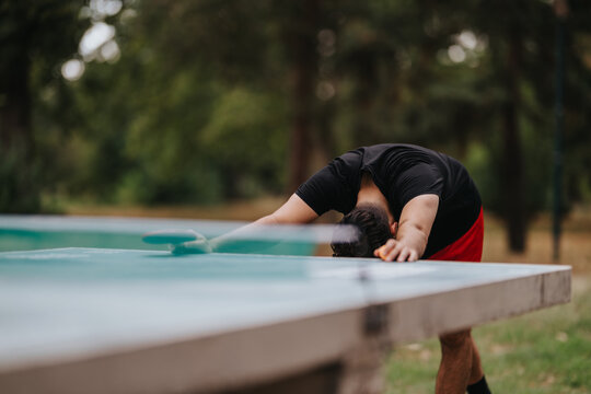 A focused athlete stretches over a park table tennis table, preparing for a rally. The outdoor setting, blurred background, and action convey fitness, concentration, and the spirit of outdoor sport. - Powered by Adobe