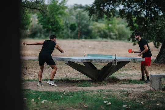 Two men in athletic wear face off at a concrete park table tennis table, trading spins under trees in a casual, energetic outdoor game.