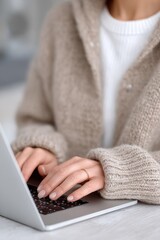 Naklejka premium Person in warm beige knit sweater typing on laptop, soft natural light and wooden desk