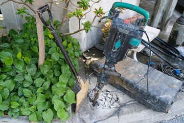 Electric jackhammer and shovel resting beside a broken concrete surface near green plants and construction tools