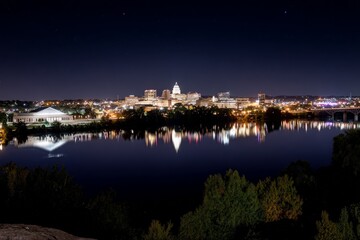 Fototapeta premium City lights glow brightly at night, modern buildings reflect on smooth river surface, dark starry sky