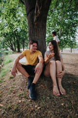 A young couple rests against a large tree in a leafy park, chatting and smiling while a friend...
