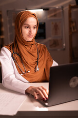 Thoughtful Muslim female doctor wearing hijab and stethoscope working in office space using laptop...
