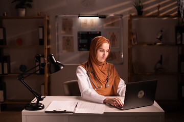 Thoughtful Muslim female doctor wearing hijab and stethoscope working in office space using laptop...