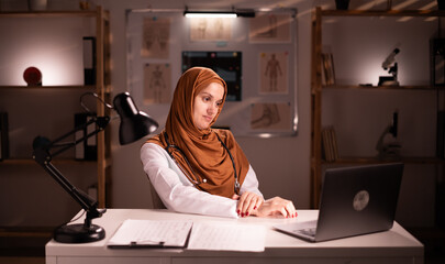 Muslim female doctor wearing hijab and stethoscope working in office using laptop computer in...