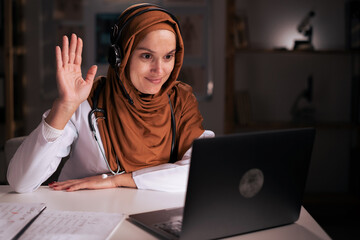 Muslim female doctor waving hand on video call looking on laptop screen, patient consulting...