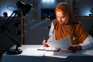 Muslim doctor woman with clipboard in office working on night shift in hospital with healthcare,...