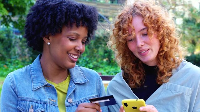  Cheerful African American and Caucasian friends stand in a city park, holding colorful smartphones and a credit card, celebrating cashless convenience, shared shopping success, and digital lifestyle