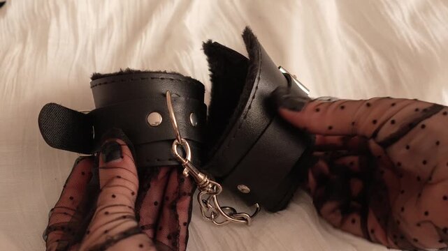 Woman in black transparent gloves holds black leather handcuffs with soft middle. BDSM erotic set. Adult toys.