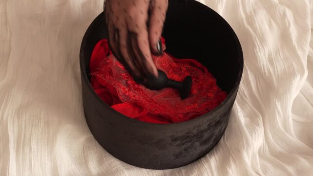 Woman in transparent glove opens black velvet box with black butt plug and red transparent lace lingerie. BDSM erotic set. Adult toys.